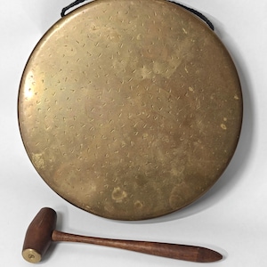 May include: A circular brass gong featuring a textured surface with small, repeating V-shaped indentations and a black cord for hanging. Below the gong lies a wooden mallet with a cylindrical head and a tapered handle, featuring a small brass inlay.