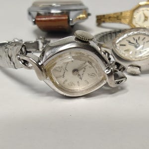 May include: A collection of four vintage wristwatches featuring various designs. One watch has a silver-tone case with a matching metal link band, while another features a gold-tone finish. A third watch includes a brown leather strap, and a fourth has a silver-tone expansion band. Each watch displays a round or octagonal face with analog hands and markers.