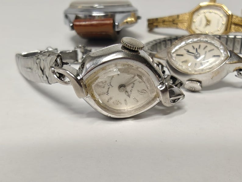 May include: A collection of four vintage wristwatches featuring various designs. One watch has a silver-tone case with a matching metal link band, while another features a gold-tone finish. A third watch includes a brown leather strap, and a fourth has a silver-tone expansion band. Each watch displays a round or octagonal face with analog hands and markers.