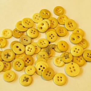 May include: A collection of approximately 30 yellow plastic buttons with two, four, or six holes. The buttons are arranged in a pile on a white surface.