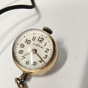 May include: A round gold-tone pendant watch with a white face, black Arabic numerals, and the word IMPERIAL printed in black. The watch features a small winding crown at the top and is attached to a thin black cord and a gold-tone metal chain.