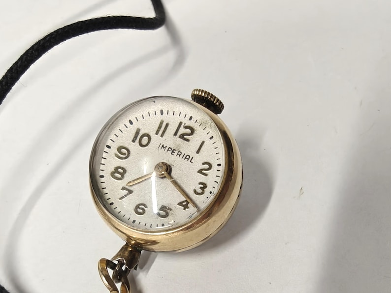 May include: A round gold-tone pendant watch with a white face, black Arabic numerals, and the word IMPERIAL printed in black. The watch features a small winding crown at the top and is attached to a thin black cord and a gold-tone metal chain.