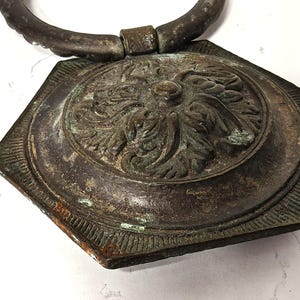 May include: A dark, weathered metal door knocker featuring a hexagonal backplate with a raised, intricate floral relief pattern. The piece has a circular ring handle attached to the top, showing signs of oxidation and a tarnished, aged patina.