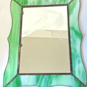 May include: A rectangular mirror with a wavy, green stained-glass frame. The frame features shades of green and white, with a dark brown metal border. The mirror is designed for wall hanging.