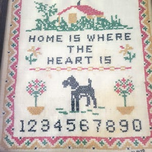 May include: Framed cross-stitch artwork with the text "HOME IS WHERE THE HEART IS." The design includes a house, dog, flowers, and the numbers 1-9. The border is a repeating pattern of pink, green, and yellow.