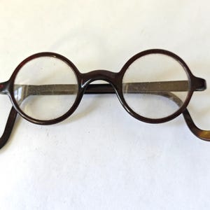 May include: Round, brown-framed eyeglasses with clear lenses. The glasses have a classic, vintage design with a bridge and temples. The frames are a dark brown color, and the lenses are circular. The temples curve down to rest behind the ears.