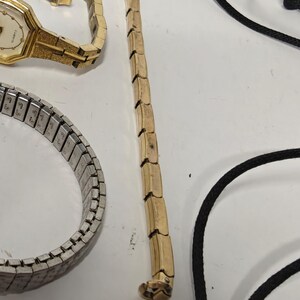 May include: A collection of jewelry items including a gold-tone quartz wristwatch with a textured link band, a silver-tone expandable metal watch band, a long gold-tone metal chain, and a black cord necklace with a simple clasp mechanism.