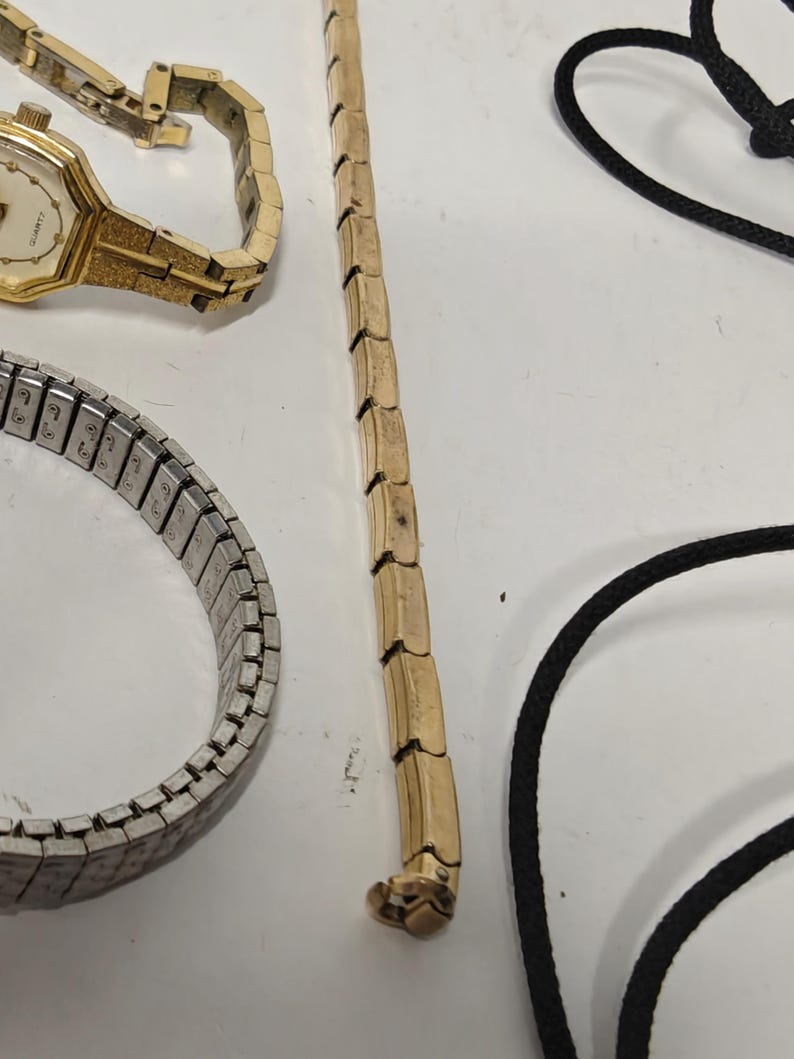 May include: A collection of jewelry items including a gold-tone quartz wristwatch with a textured link band, a silver-tone expandable metal watch band, a long gold-tone metal chain, and a black cord necklace with a simple clasp mechanism.