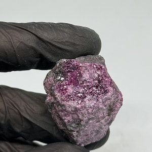 Peut inclure: Spécimen minéral naturel brut, avec un extérieur gris foncé et un intérieur cristallin violet profond. Les cristaux étincellent à l'intérieur, contrastant avec la surface extérieure plus sombre. Le spécimen est tenu par une main gantée.