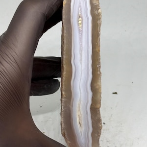May include: A vertical slice of agate stone featuring a central band of translucent white and light blue layers, surrounded by thicker, semi-translucent tan and brown outer edges with a rough, natural texture along the vertical sides.