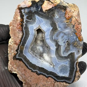 May include: A geode slice featuring a rough, tan-coloured outer crust and a polished interior. The centre displays concentric bands of light blue and dark grey agate surrounding a hollow cavity filled with small, sparkling white quartz crystals.