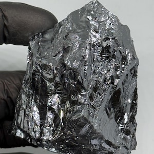 May include: A large, dark grey silicon crystal with an irregular shape. The surface is highly reflective, with many facets catching the light. The crystal is held by a gloved hand, showcasing its size and texture. It appears to be a raw, natural mineral specimen.