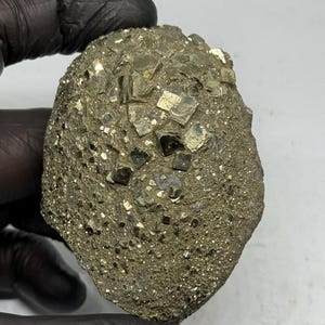 May include: A raw pyrite mineral specimen featuring a cluster of metallic gold-colored cubic crystals embedded in a textured, earthy matrix. The surface displays a mix of larger, distinct geometric cubes and smaller, granular crystalline formations.