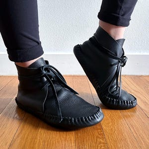 May include: Black leather ankle boots with a moccasin-style design. The boots feature a rounded toe, stitched detailing, and laces. The boots are worn with black pants, and the image is taken on a wooden floor.