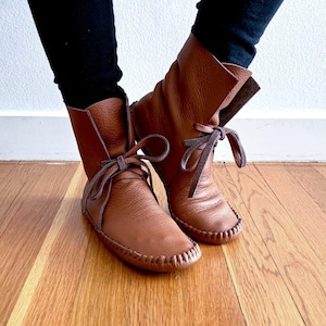 May include: A pair of mid-calf brown leather boots featuring a soft, flexible construction with a moccasin-style stitched toe seam. The boots include a wrap-around leather lace tie closure at the front, creating a gathered look at the ankle.