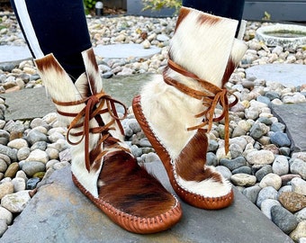 May include: Handmade ankle boots with a cowhide pattern in brown and white. The boots have a leather sole and are laced up with brown leather laces. The boots are shown outdoors on a stone surface.