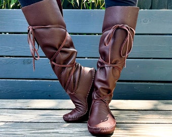 May include: Brown leather knee-high boots with laces wrapped around the legs. The boots have a moccasin-style toe and are worn on a wooden surface.