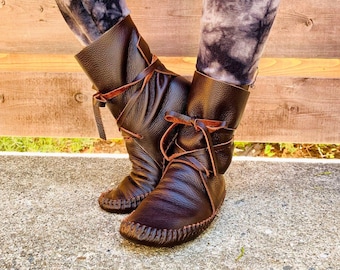 May include: A pair of dark brown leather ankle boots featuring a soft, flexible construction with visible moccasin-style stitching around the toe and a thin leather lace that wraps around the ankle and ties in a bow at the front.