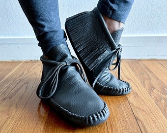 May include: A pair of black leather ankle boots with fringe detailing and laces. The boots are worn on a wooden floor.