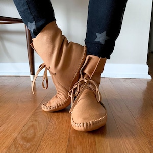 May include: A pair of tan leather ankle boots with laces. The boots have a pointed toe and a stitched seam around the top of the boot.
