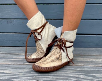 May include: Ankle-high moccasins featuring a combination of white and brown speckled animal hide. The footwear includes brown leather laces that tie at the front and a dark brown leather sole with visible whipstitch detailing along the edges.