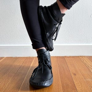 May include: Black leather ankle boots with a moccasin-style design. The boots feature a rounded toe, stitched detailing, and leather laces. The boots are worn with black pants, and the background is a light-colored wall and wooden floor.