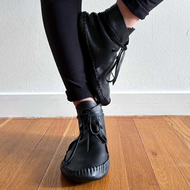 May include: Black leather ankle boots with a moccasin-style design. The boots feature a rounded toe, stitched detailing, and leather laces. The boots are worn with black pants, and the background is a light-colored wall and wooden floor.