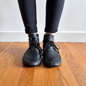 May include: Black leather ankle boots with a moccasin-style design. The boots have a rounded toe, stitched sole, and lace-up closure. The boots are worn with black pants, and the background is a wooden floor and a white wall.