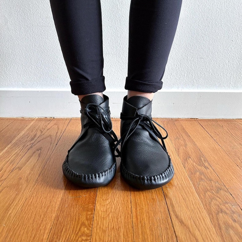 May include: Black leather ankle boots with a moccasin-style design. The boots have a rounded toe, stitched sole, and lace-up closure. The boots are worn with black pants, and the background is a wooden floor and a white wall.