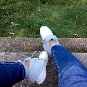 May include: White moccasin-style shoes with light blue suede cuffs and laces. The shoes are worn with blue jeans. The image is taken from a high angle, with a background of green grass and a concrete step.