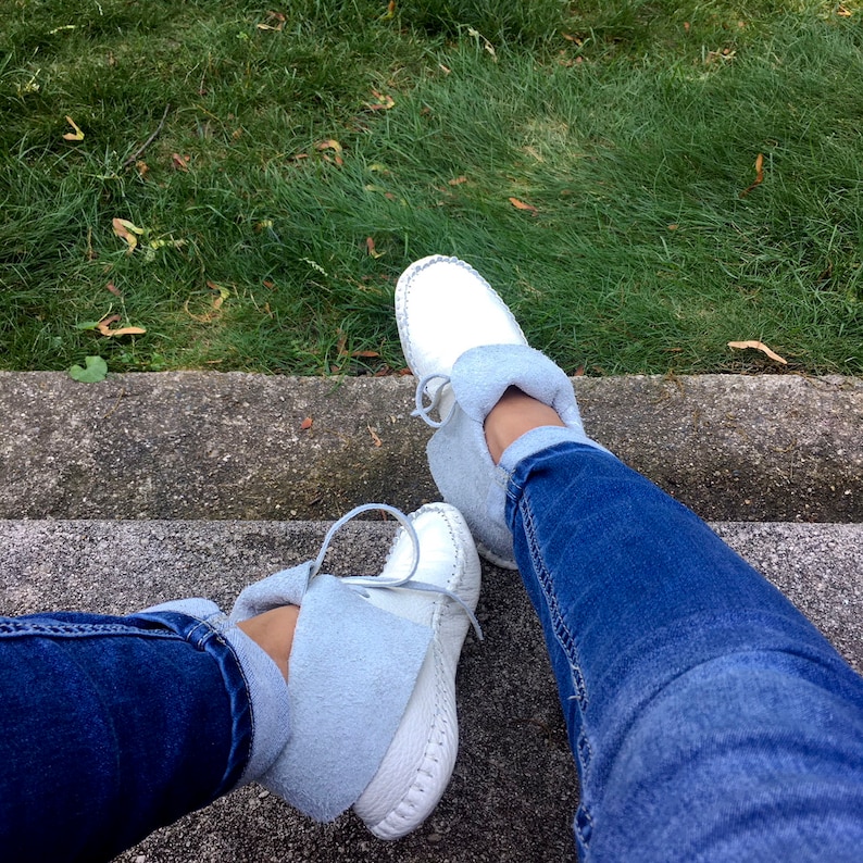 May include: White moccasin-style shoes with light blue suede cuffs and laces. The shoes are worn with blue jeans. The image is taken from a high angle, with a background of green grass and a concrete step.