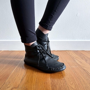 May include: Black leather ankle boots with a moccasin-style toe and lace-up closure. The boots have a textured finish and are worn with black leggings. The boots are on a wooden floor.