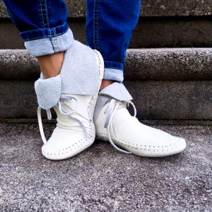 May include: White leather moccasin boots with gray suede cuffs and laces. The boots have a rounded toe and are stitched along the seams. The cuffs are folded over, revealing a soft, gray interior. The boots are worn with blue jeans.