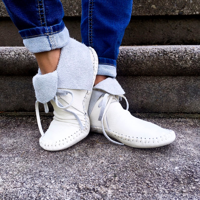 May include: White leather moccasin boots with gray suede cuffs and laces. The boots have a rounded toe and are stitched along the seams. The cuffs are folded over, revealing a soft, gray interior. The boots are worn with blue jeans.