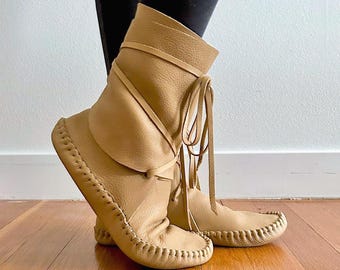 May include: A pair of tan leather mid-calf boots featuring a soft, slouchy upper, a rounded toe, and visible whipstitch detailing along the sole. The boots are secured with thin leather laces that wrap around the ankle and tie in the front.