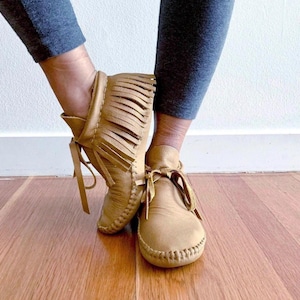 May include: A pair of tan leather moccasins featuring a rounded toe, visible whipstitch detailing along the sole, and a decorative fringe panel on the outer side of the ankle. The footwear includes thin leather laces tied in a bow at the front.