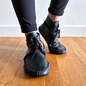 May include: Black leather ankle boots with a moccasin-style design. The boots feature a rounded toe, stitched detailing, and a lace-up closure. The boots are worn with black pants, and the image is taken on a wooden floor.