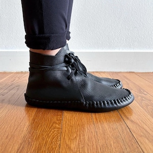 May include: Black leather ankle boots with a moccasin-style toe and stitched detailing. The boots have a lace-up closure and are worn with black pants. The boots are on a hardwood floor.