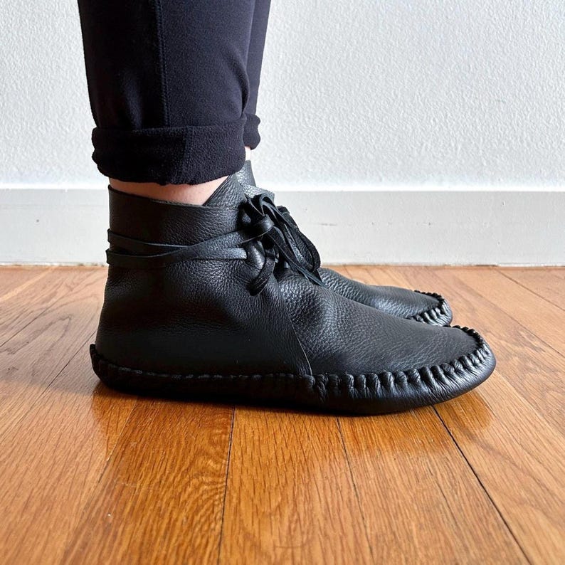 May include: Black leather ankle boots with a moccasin-style toe and stitched detailing. The boots have a lace-up closure and are worn with black pants. The boots are on a hardwood floor.