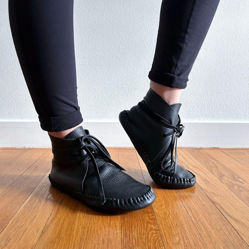 May include: Black leather ankle boots with a moccasin-style design. The boots feature a rounded toe, stitched detailing, and laces. The boots are worn with black pants, and the image is taken on a wooden floor.