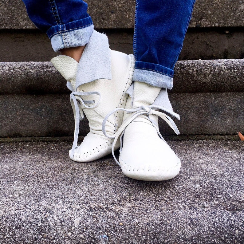 May include: Cream-colored leather moccasin boots with gray laces tied in bows. The boots have a folded-over cuff and are worn with blue jeans. The boots are on a concrete surface.