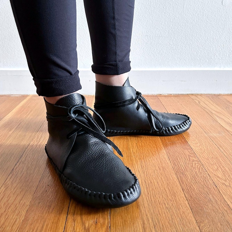 May include: Black leather ankle boots with a moccasin-style design. The boots feature a rounded toe, stitched detailing, and a lace-up closure. The boots are worn with black pants on a wooden floor.