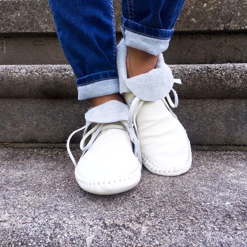 May include: A pair of white leather moccasin-style shoes with light gray suede collars and laces. The shoes are worn with blue jeans with the cuffs rolled up, and are sitting on a concrete surface.