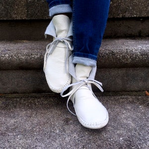 May include: White leather moccasin boots with gray laces and stitching. The boots are tied with gray laces and are paired with blue jeans. The boots have a soft, folded-over cuff.