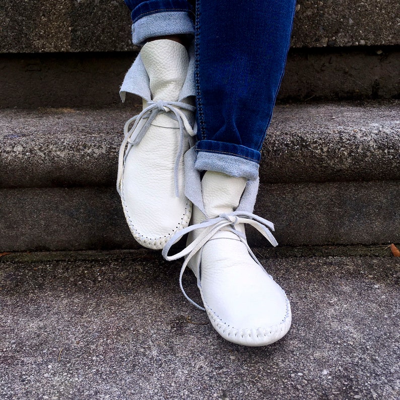 May include: White leather moccasin boots with gray laces and stitching. The boots are tied with gray laces and are paired with blue jeans. The boots have a soft, folded-over cuff.