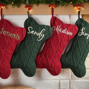 May include: Four personalized Christmas stockings in red and green, hanging from a wooden mantel. Each stocking is cable-knit and embroidered with a name: Genesis, Sandy, Madison, and Simone. Festive holiday decor.
