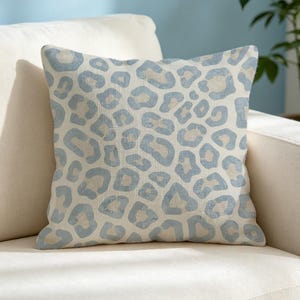 May include: A square throw pillow featuring a light blue and off-white leopard print pattern. The fabric has a visible woven texture, and the design consists of repeating, irregular animal spots distributed evenly across the entire surface.