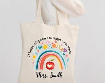 May include: Cream tote bag with a rainbow design, an apple, and the text "It Takes a Big Heart to Shape Little Minds" and "Miss. Smith". The bag has long handles and is made of a plain material.