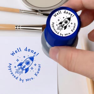 May include: A blue and white stamp with the words "Well done! Approved by Mrs. Kumar" encircling a rocket ship illustration. The stamp is held over a white paper with a matching stamped image. Paint brushes and a mirror are in the background.