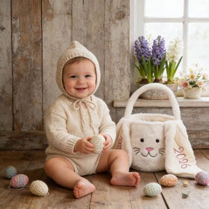 Può includere: Un bambino con un pagliaccetto e un cappuccio di maglia color crema, che tiene in mano un uovo di maglia. Un cestino a forma di coniglio di peluche e diverse uova decorative sono nelle vicinanze. Il cestino ha ricamato l'anno "2026".